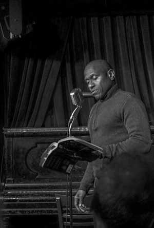 Adam Smyer in a high-necked sweater, in front of a microphone, reading from his book 'Knucklehead'